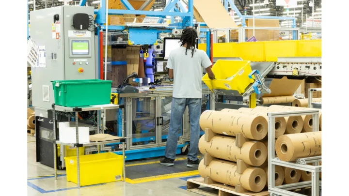 An operator at an Amazon automated paper-packaging station with paper rolls and forming equipment.