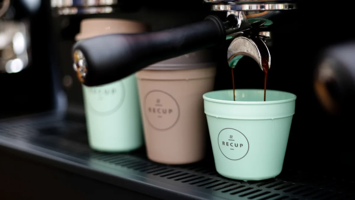 Three RECUP deposit cups beside an espresso machine, with coffee pouring into the nearest cup
