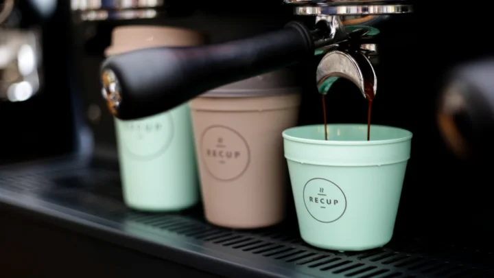 Three RECUP deposit cups beside an espresso machine, with coffee pouring into the nearest cup