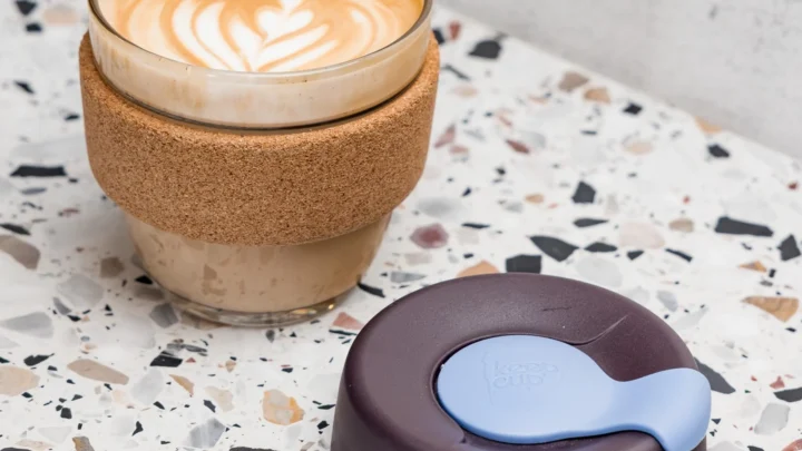 Coffee in a reusable glass cup with a cork band and its separate lid on a terrazzo tabletop