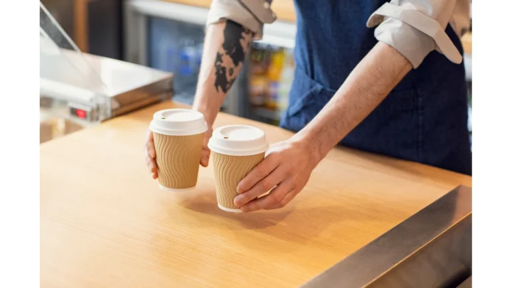 Two brown paper takeaway cups with white lids being placed on a cafe counter.