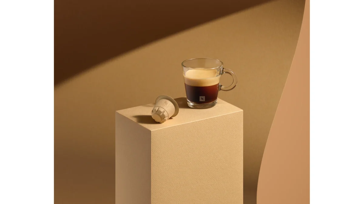 A paper-based Nespresso capsule beside an espresso in a glass cup on a beige display block.