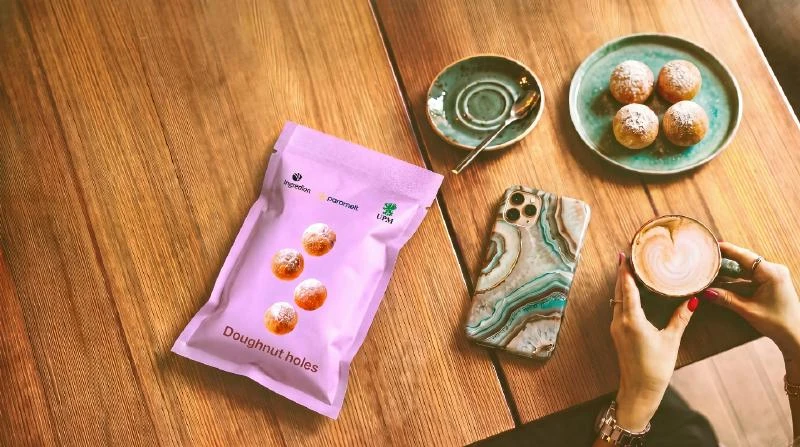 A pink paper pouch carrying UPM, Ingredion and Paramelt names on a table beside doughnuts and cups.